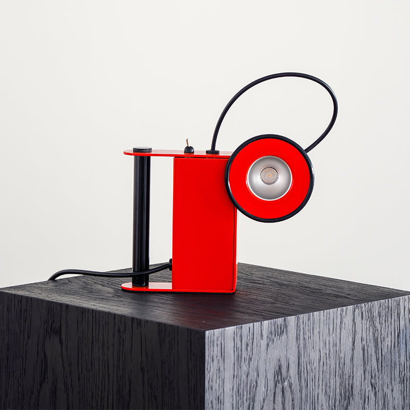 A table lamp with a red shade and black base, featuring a silver bulb and a black cord, placed on a grey surface.