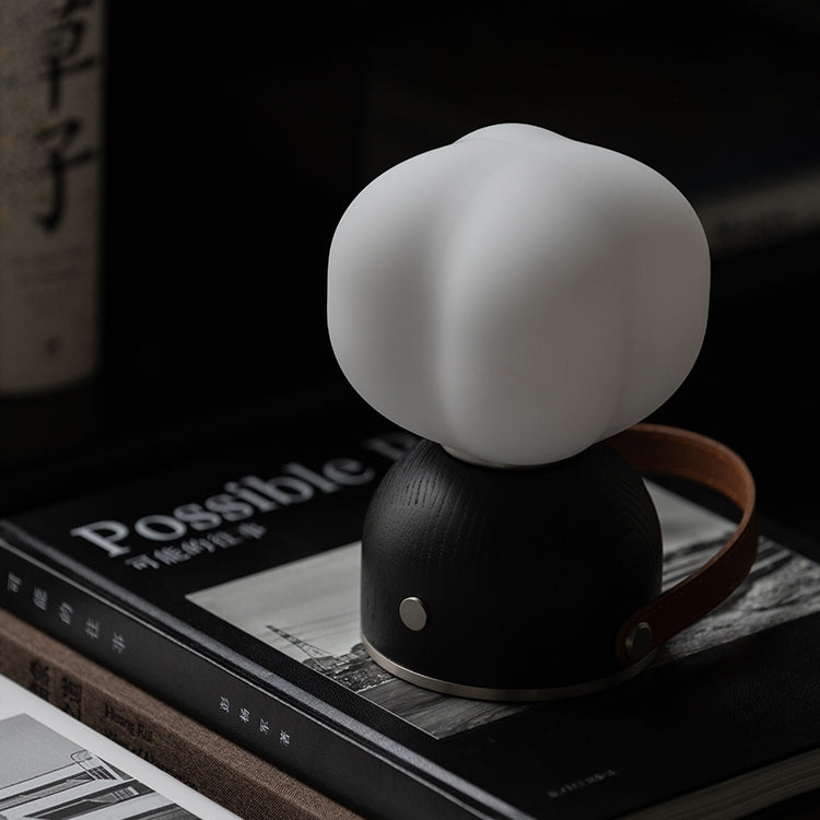 A table lamp with a white shade and black base, placed on a surface with a book in the background.