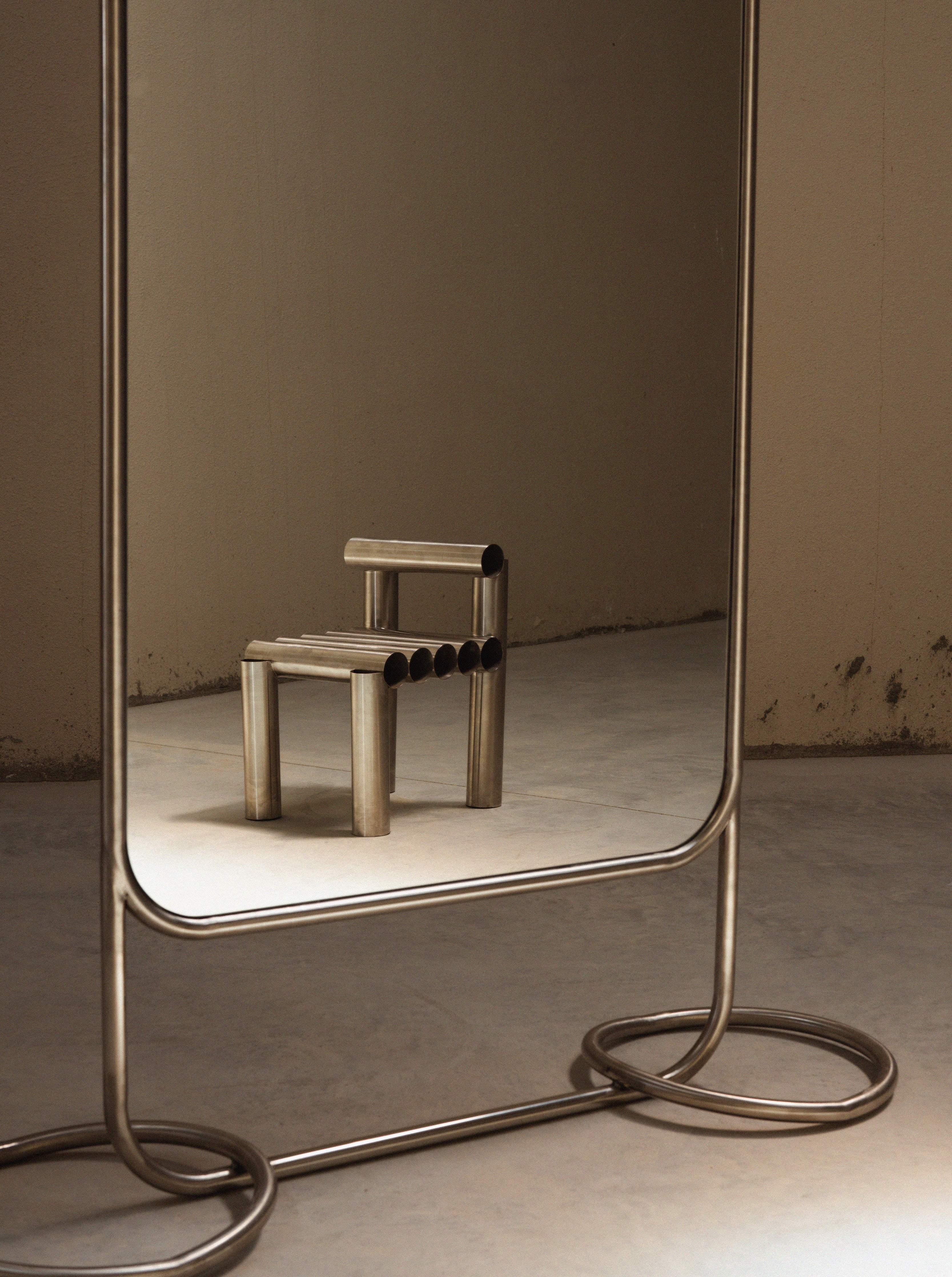 Metallic chair reflected in a standing mirror against a plain wall.
