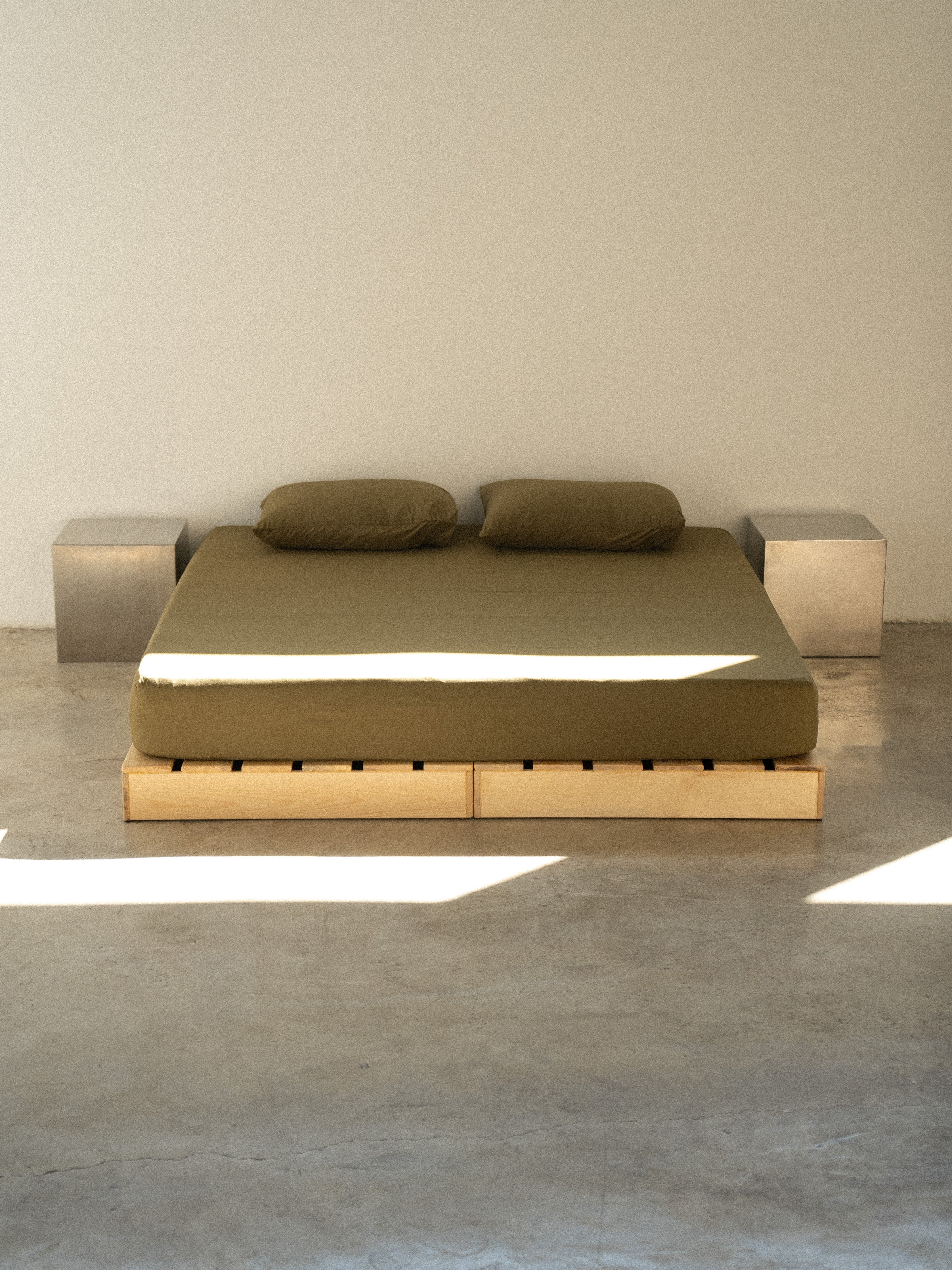 A modern bed with a beige mattress platform and green bedding, flanked by two gray nightstands, set against a white wall on a concrete floor.
