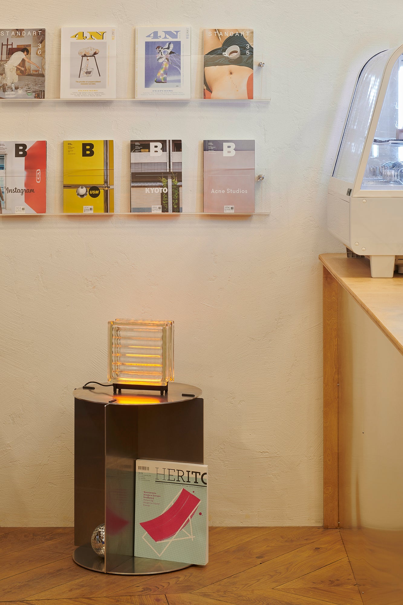 Display of books on a shelf against a beige wall with a small table and lamp in the foreground.