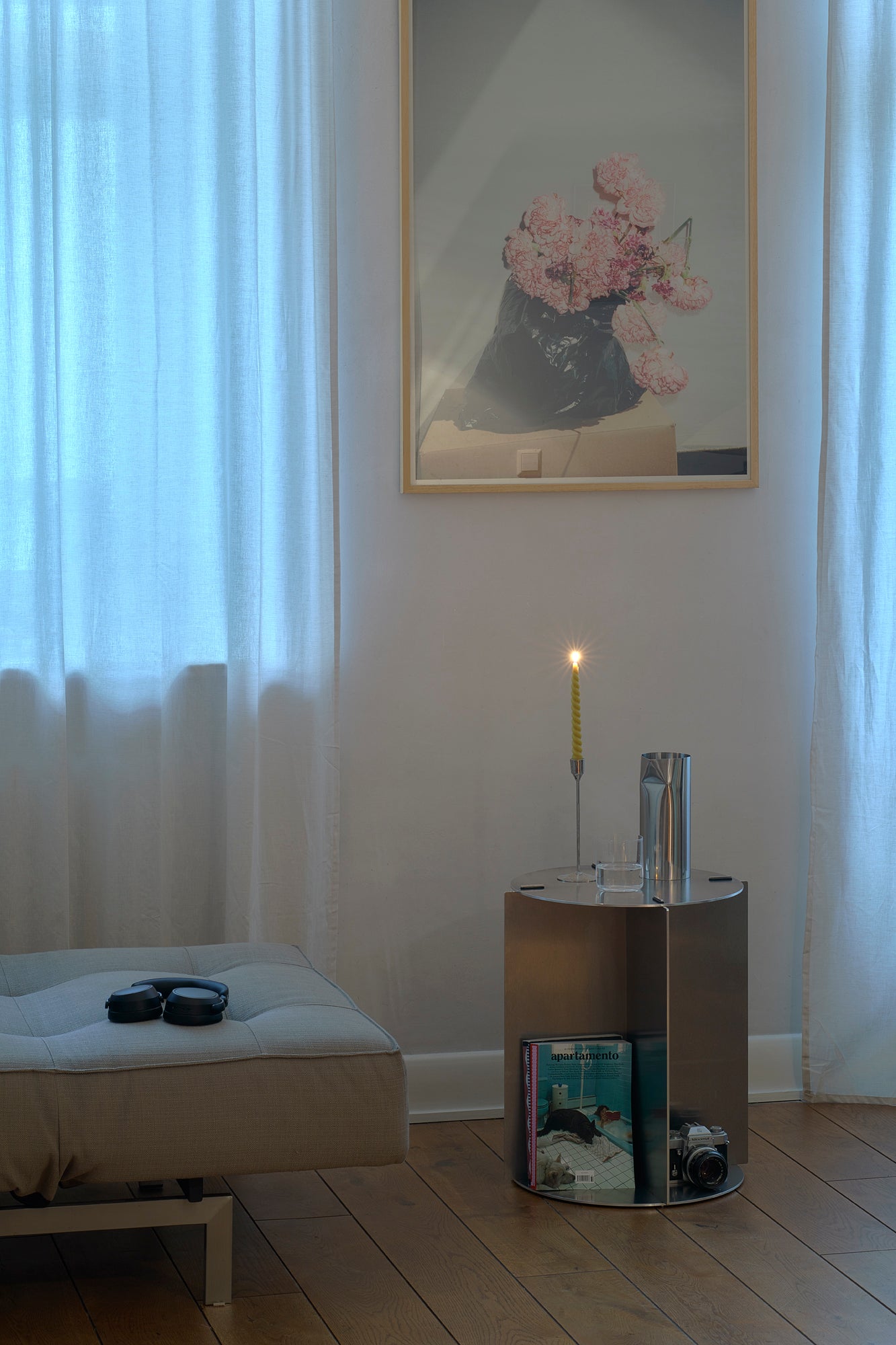 Silver cylindrical table with a candle, books, and headphones in a room with white curtains and a framed picture of flowers.