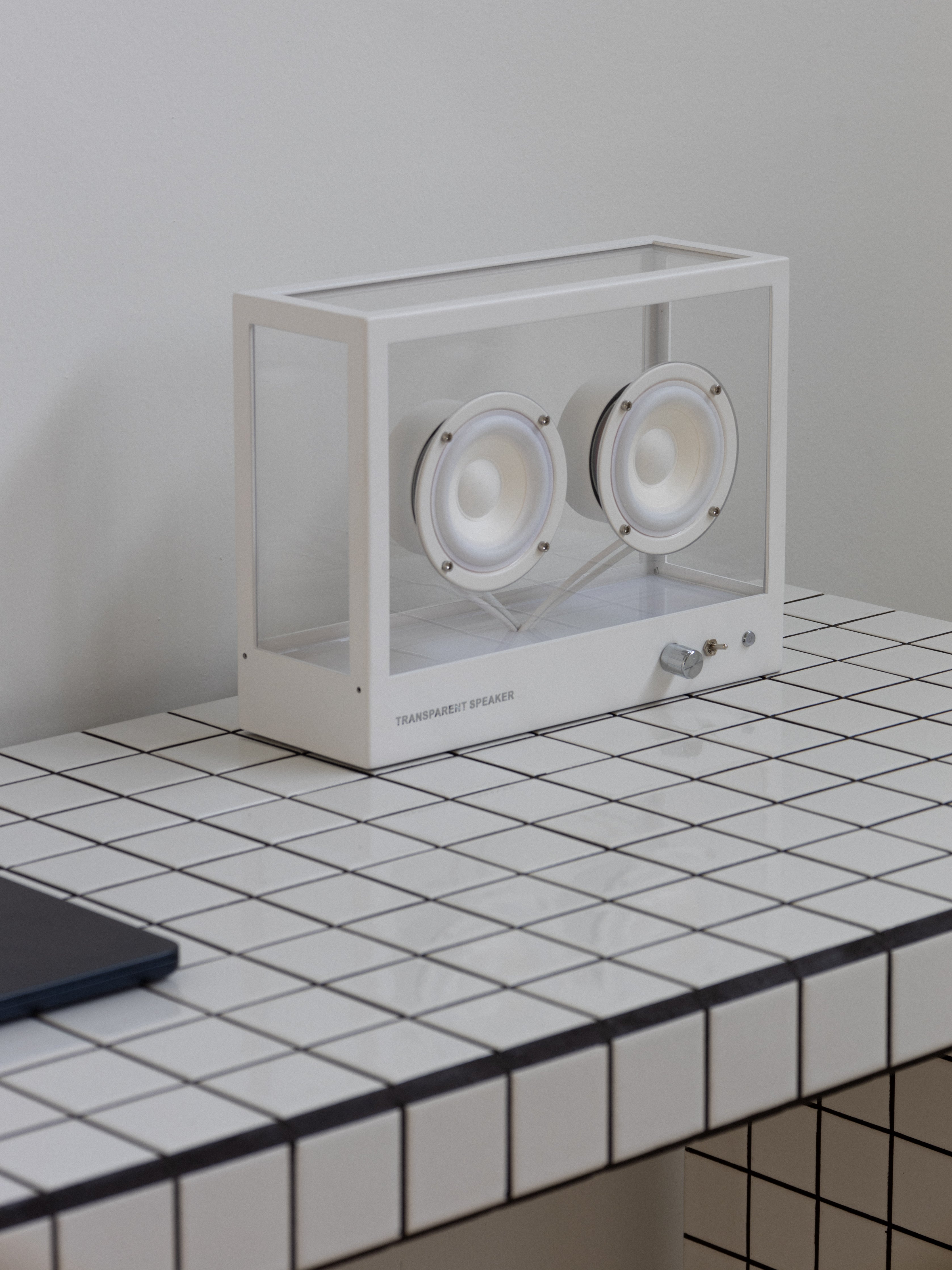 A transparent sound speaker with a white frame and visible internal components, placed on a tiled surface.