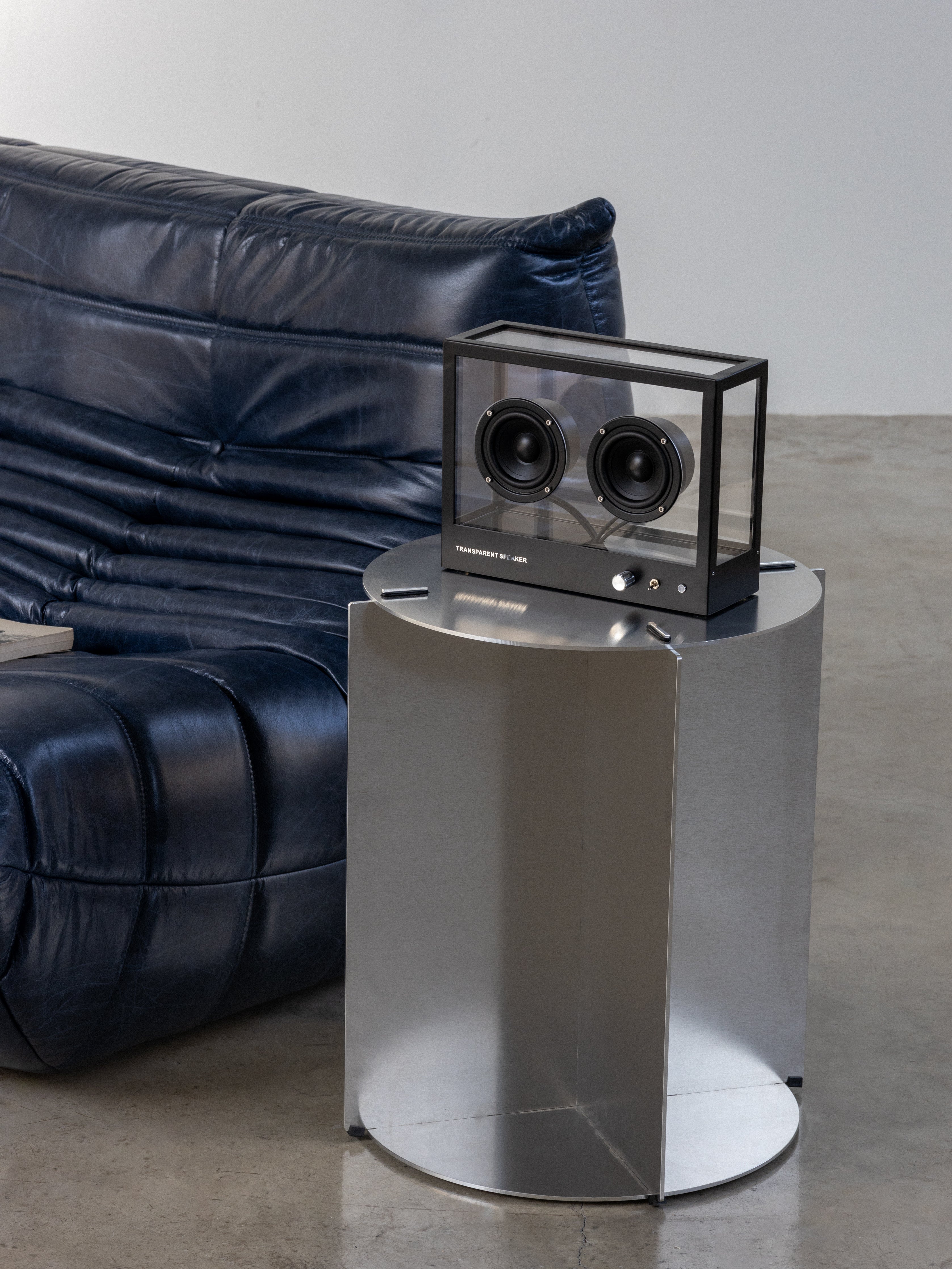Modern black speaker on a cylindrical silver stand with a blue sofa in the background.