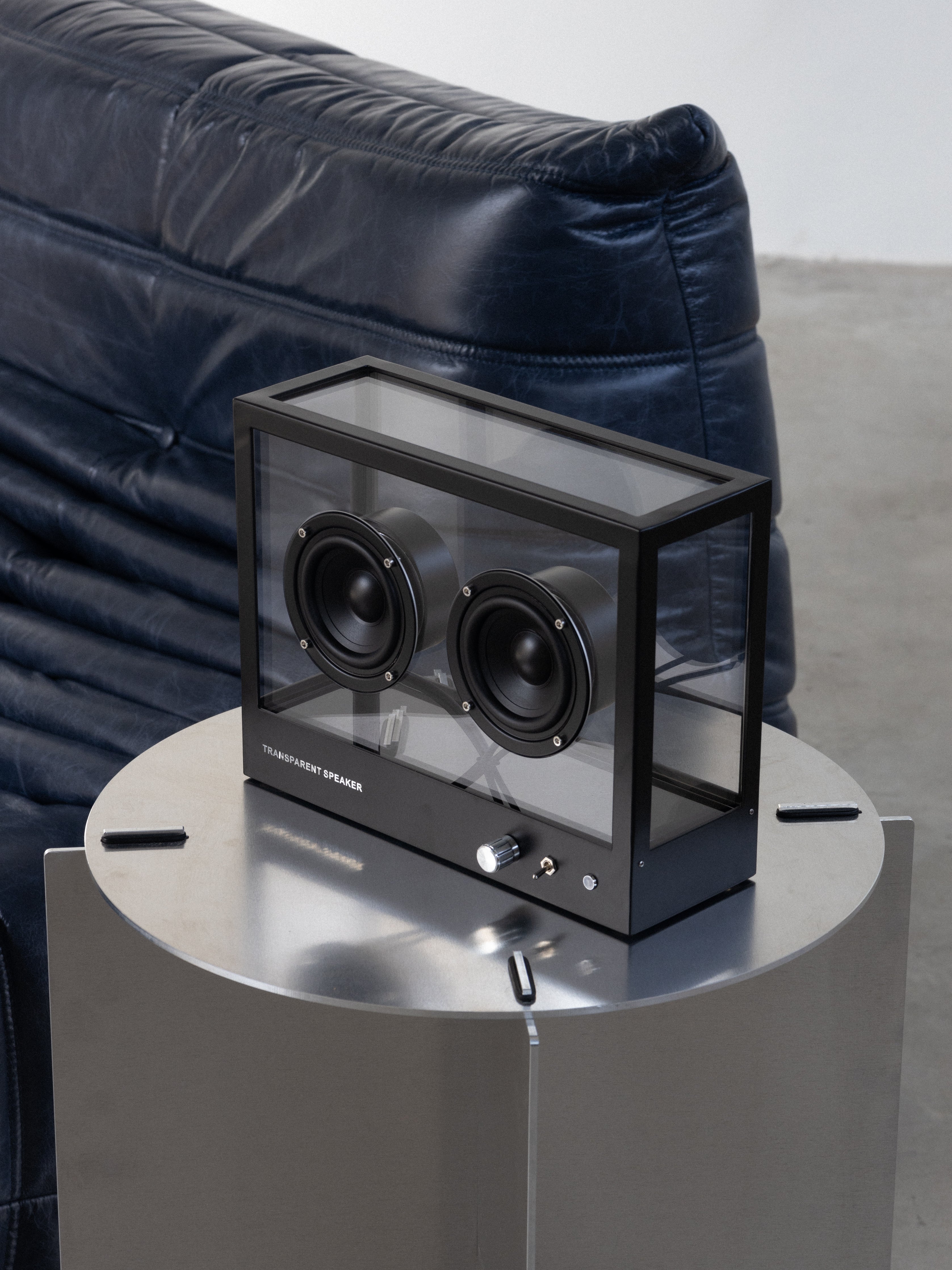 Black rectangular speaker on a metallic stand with a blue sofa in the background