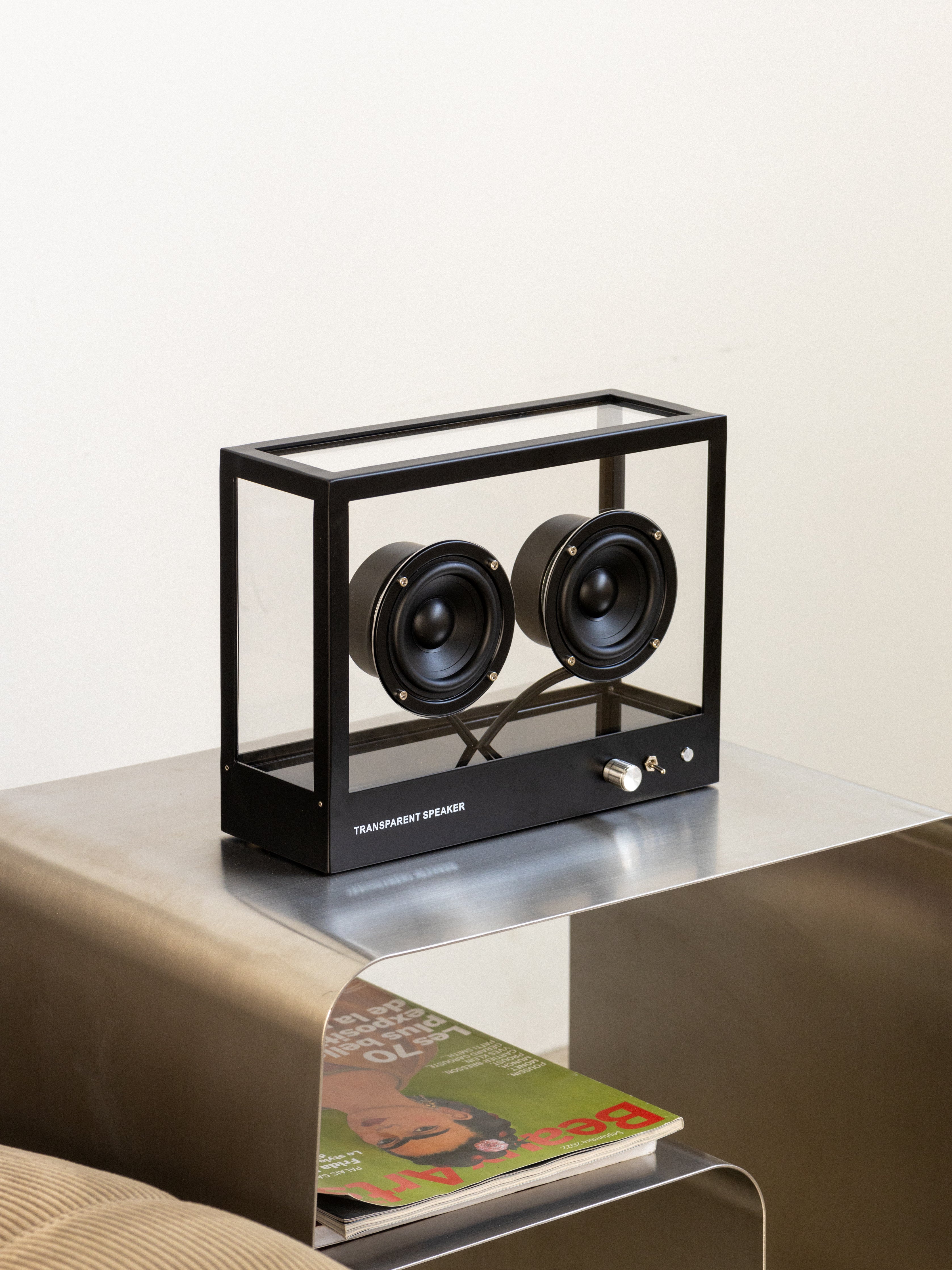 Two black speakers in a transparent black box on a reflective surface with a magazine underneath.