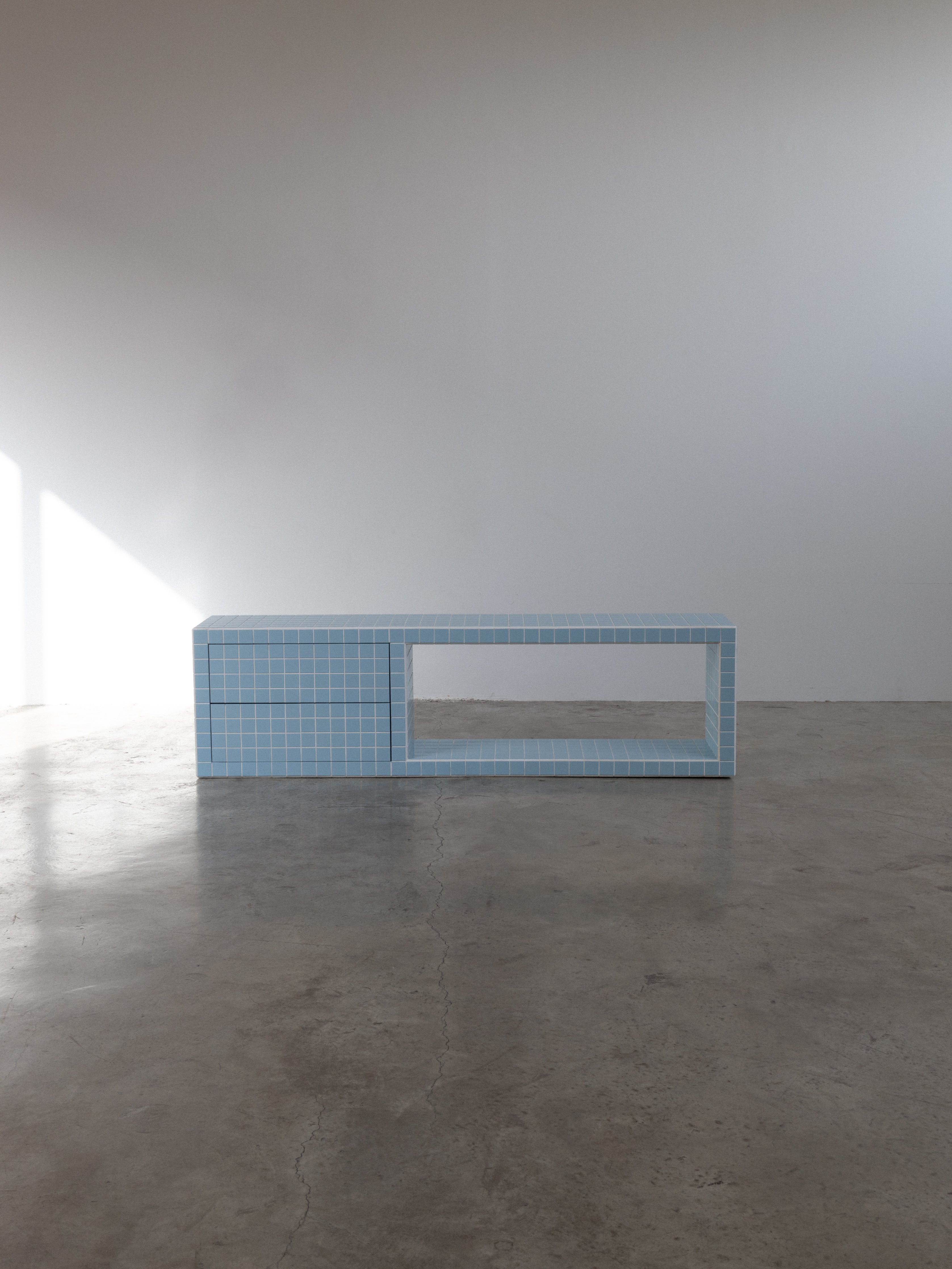 Tiled TV unit in light blue on a concrete floor, featuring a drawer and an open shelf, ideal for modern interiors.