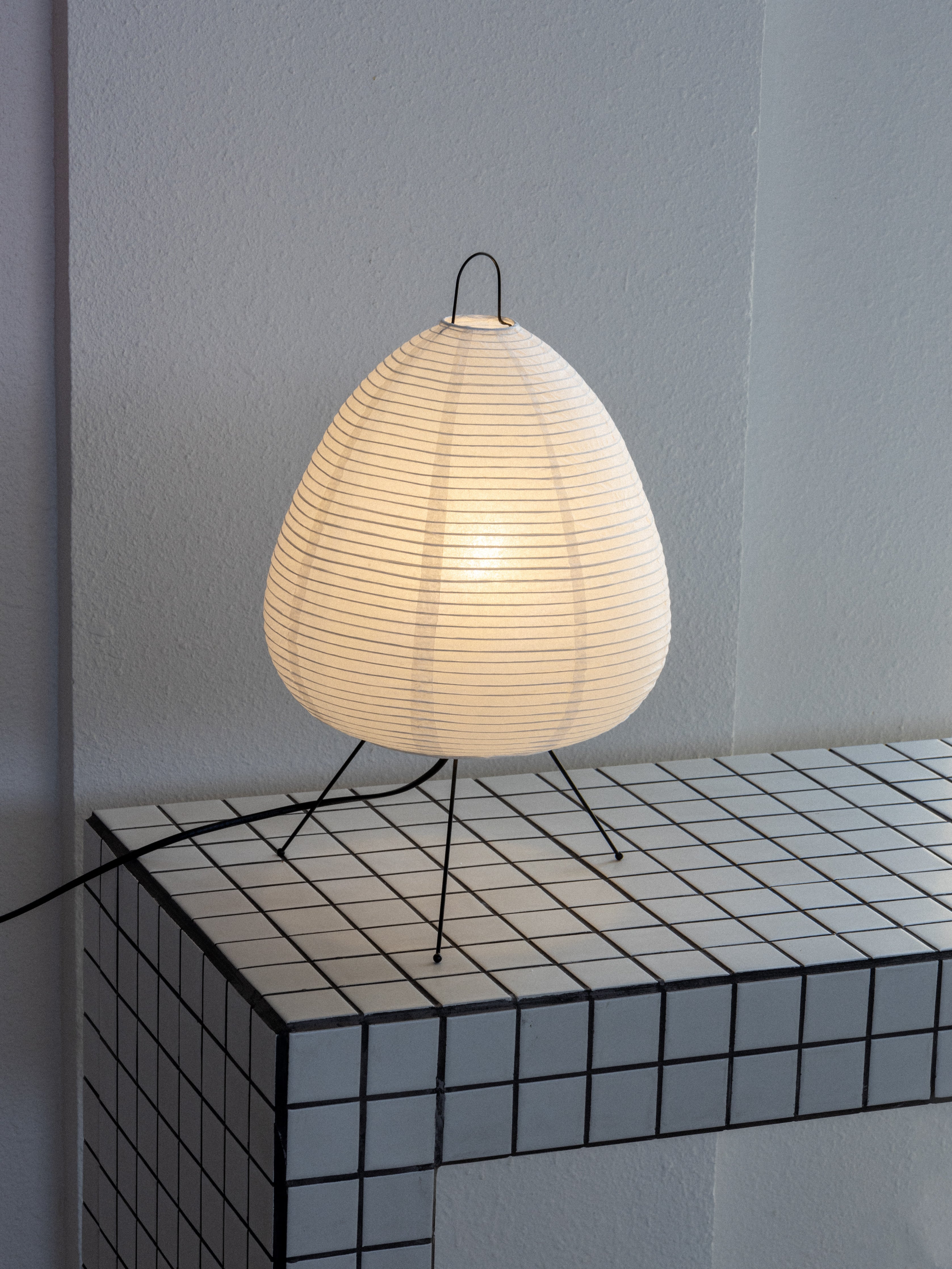 Akari 1A table lamp with a white, spherical shade on a black metal stand, illuminated, placed against a wall on a geometric patterned surface.