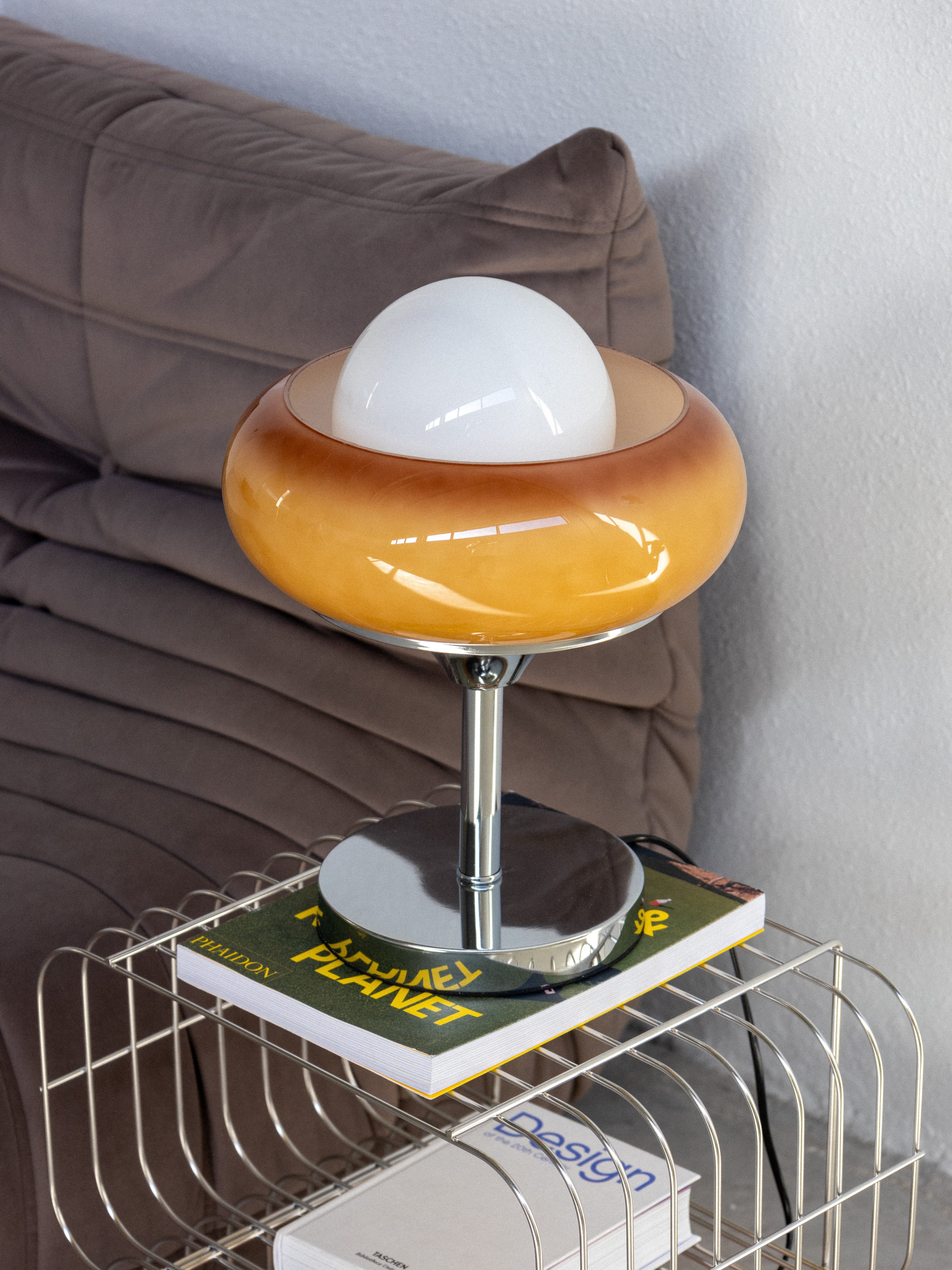 A vintage glass table lamp with a brown and white color scheme, featuring a globe-shaped shade, placed on a metal magazine rack next to a couch.