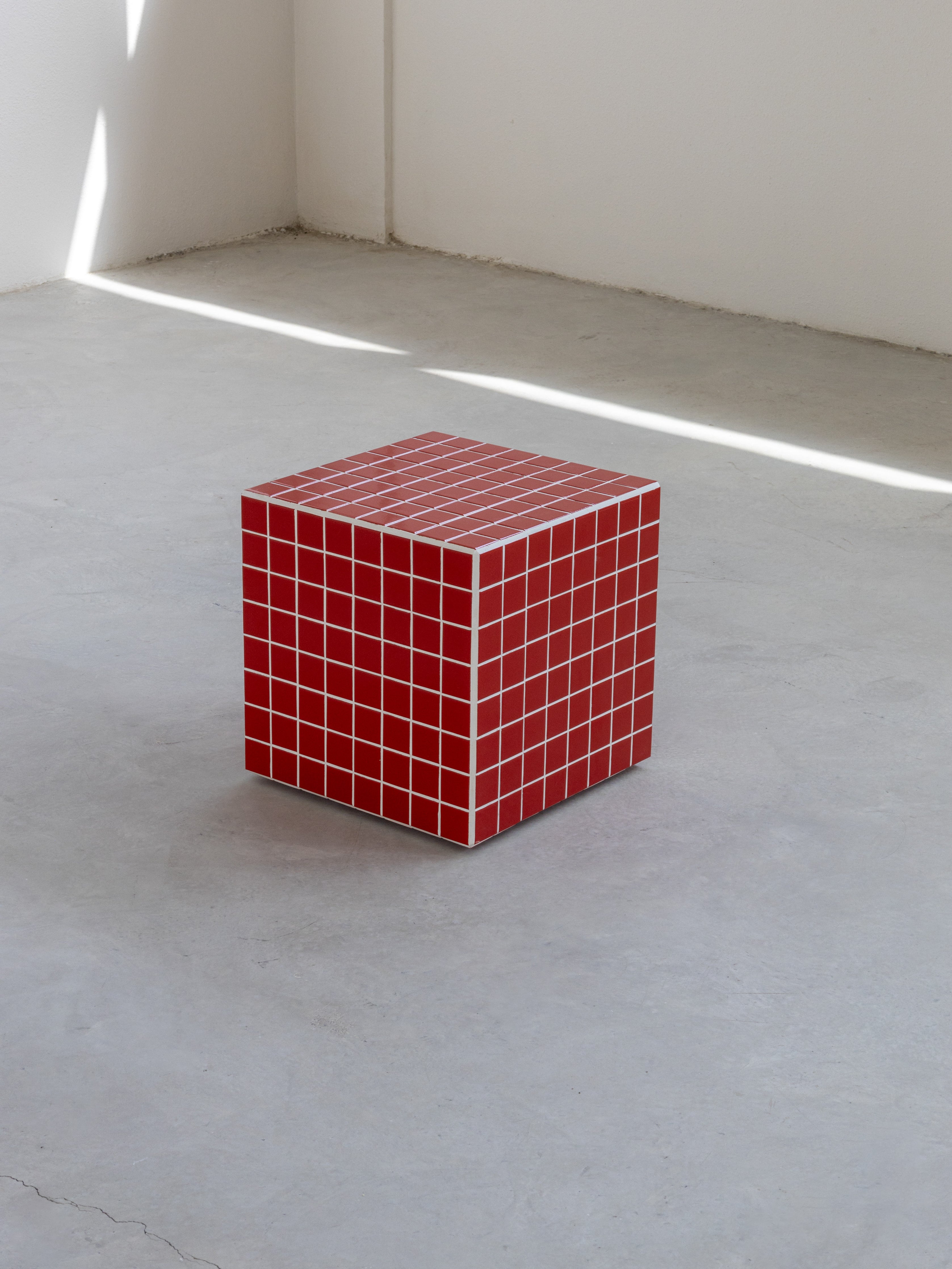 A cube-shaped table with a red and white tiled pattern, placed on a grey floor with a light source creating a shadow on the wall.