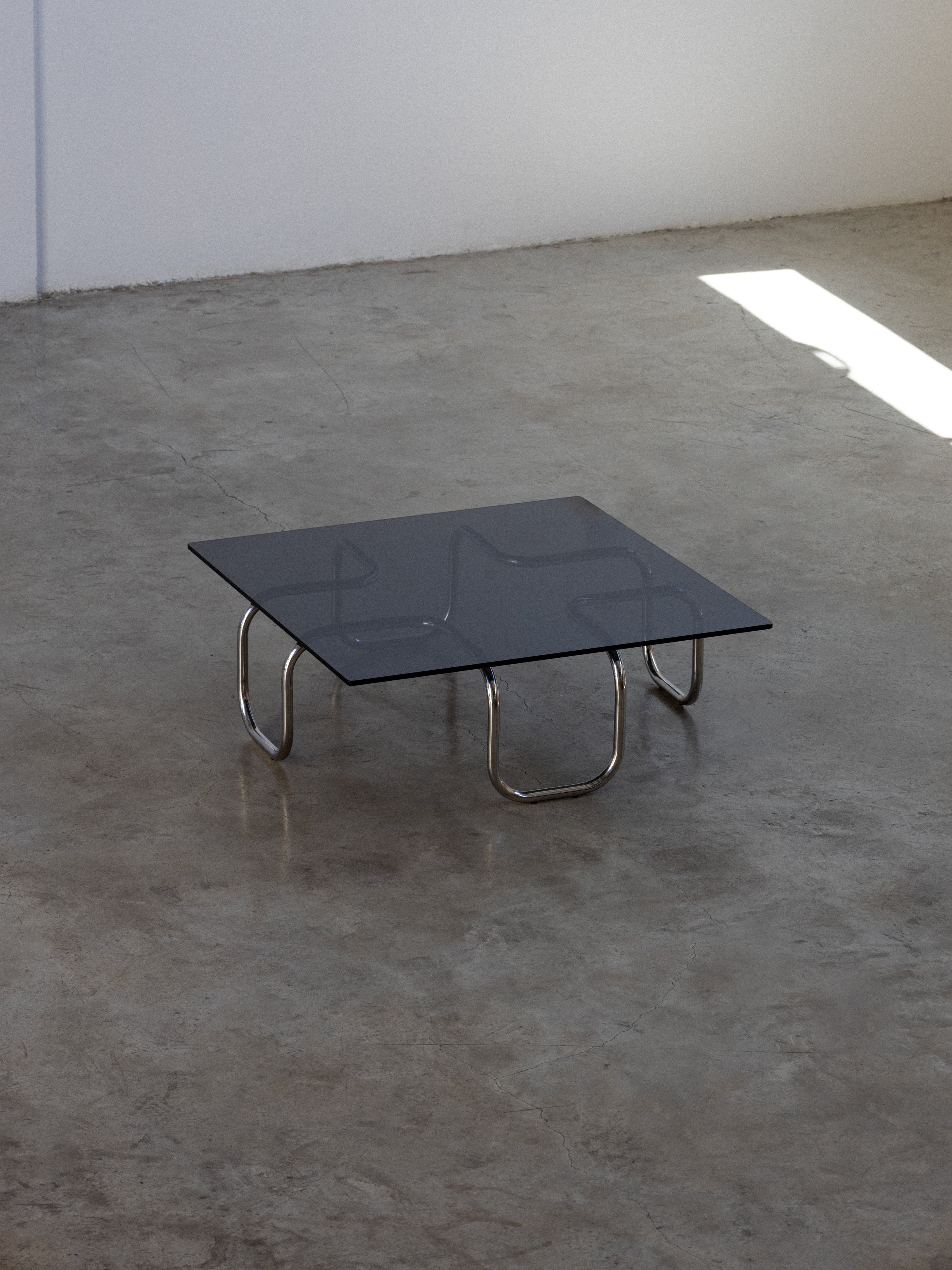 A square black glass tabletop coffee table with metal legs, placed on a concrete floor.