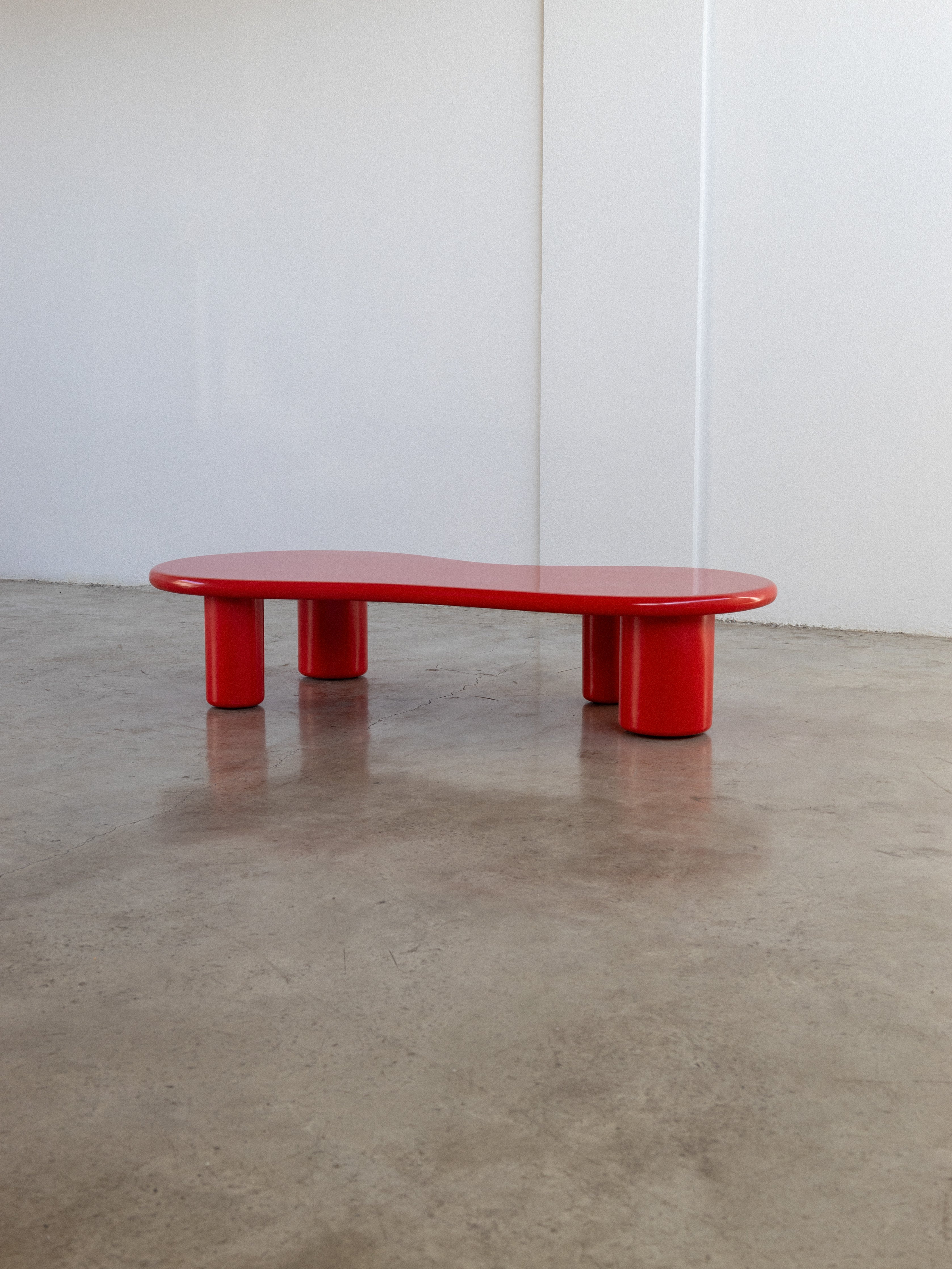 A red kidney-shaped coffee table with a black base, placed on a concrete floor against a white wall.