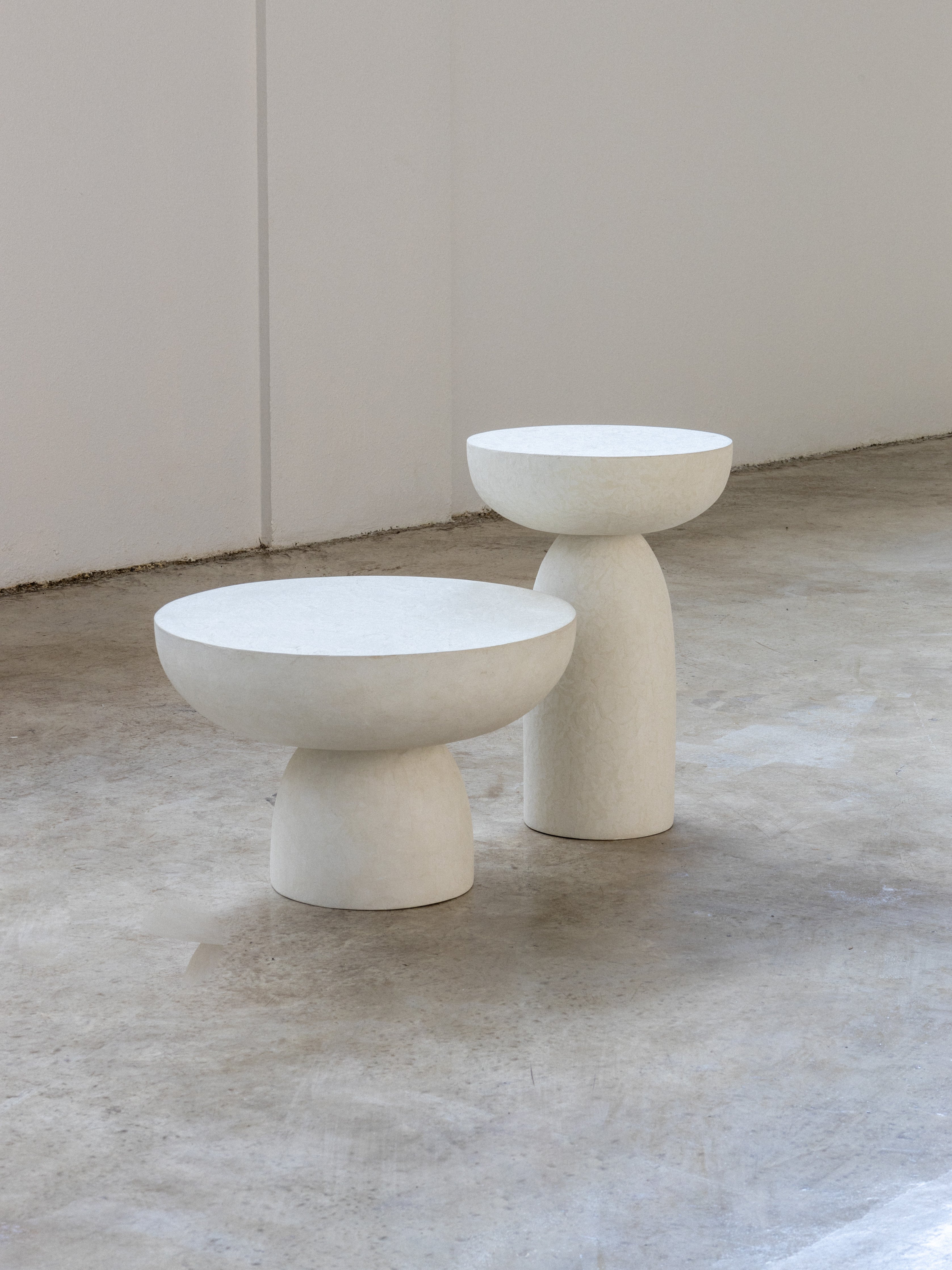 Two round concrete tables with white surfaces and beige legs placed on a grey floor.