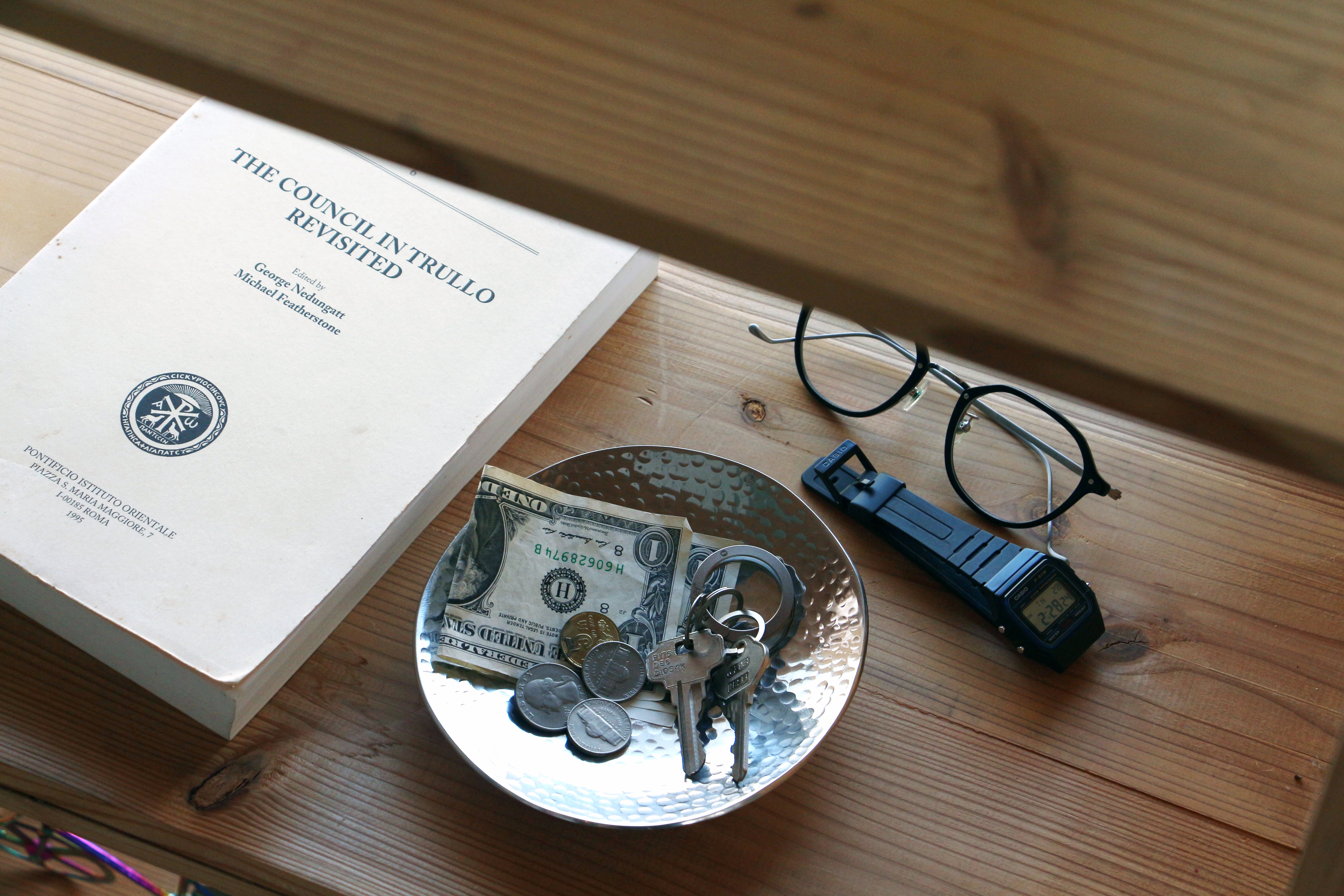 Flat lay of an Aluminum Tray holding cash and coins, with a book and glasses on a wooden table.