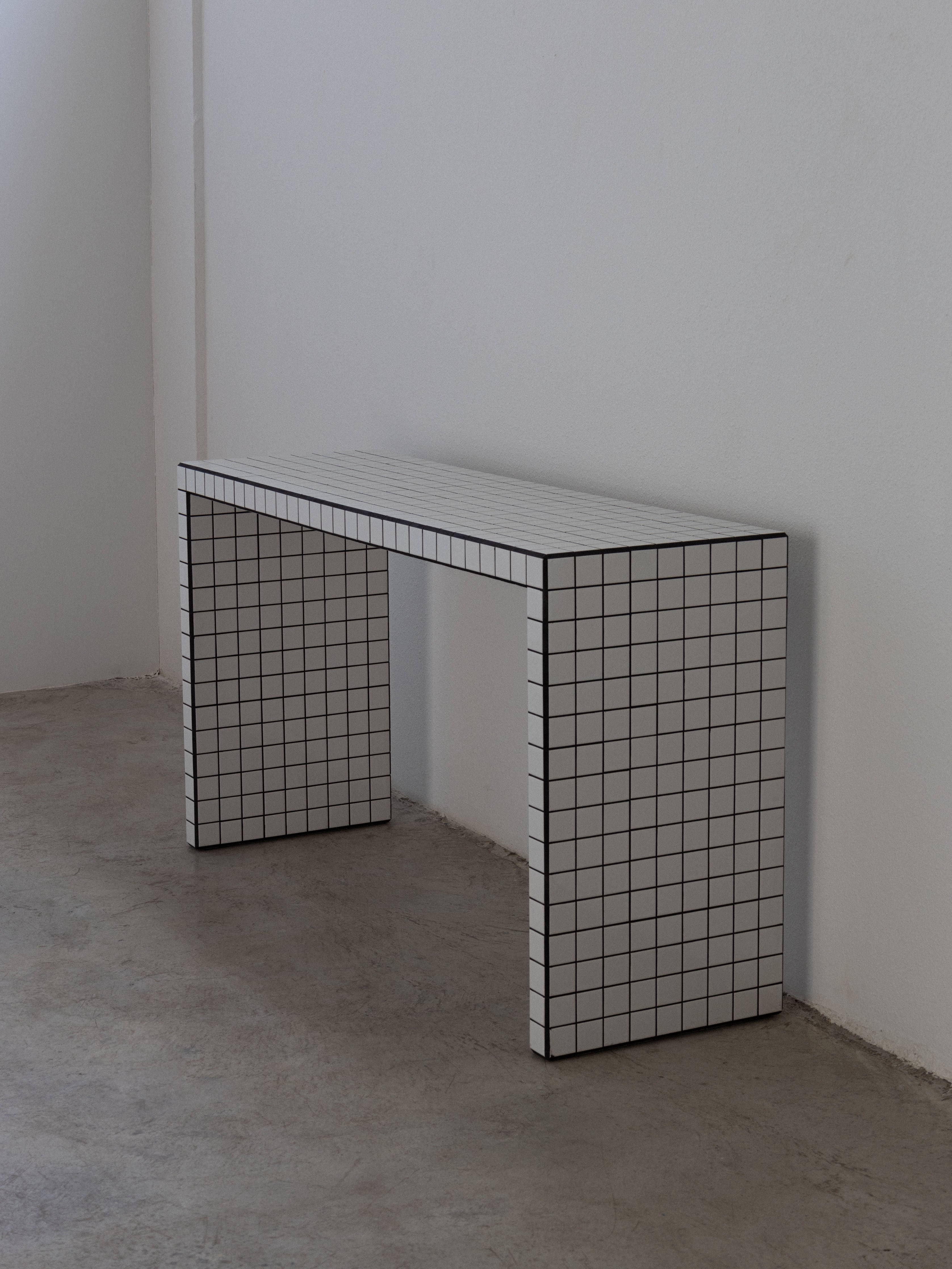 A slim console table with a white tile top and black grout lines, supported by a black frame, positioned against a wall on a neutral background.