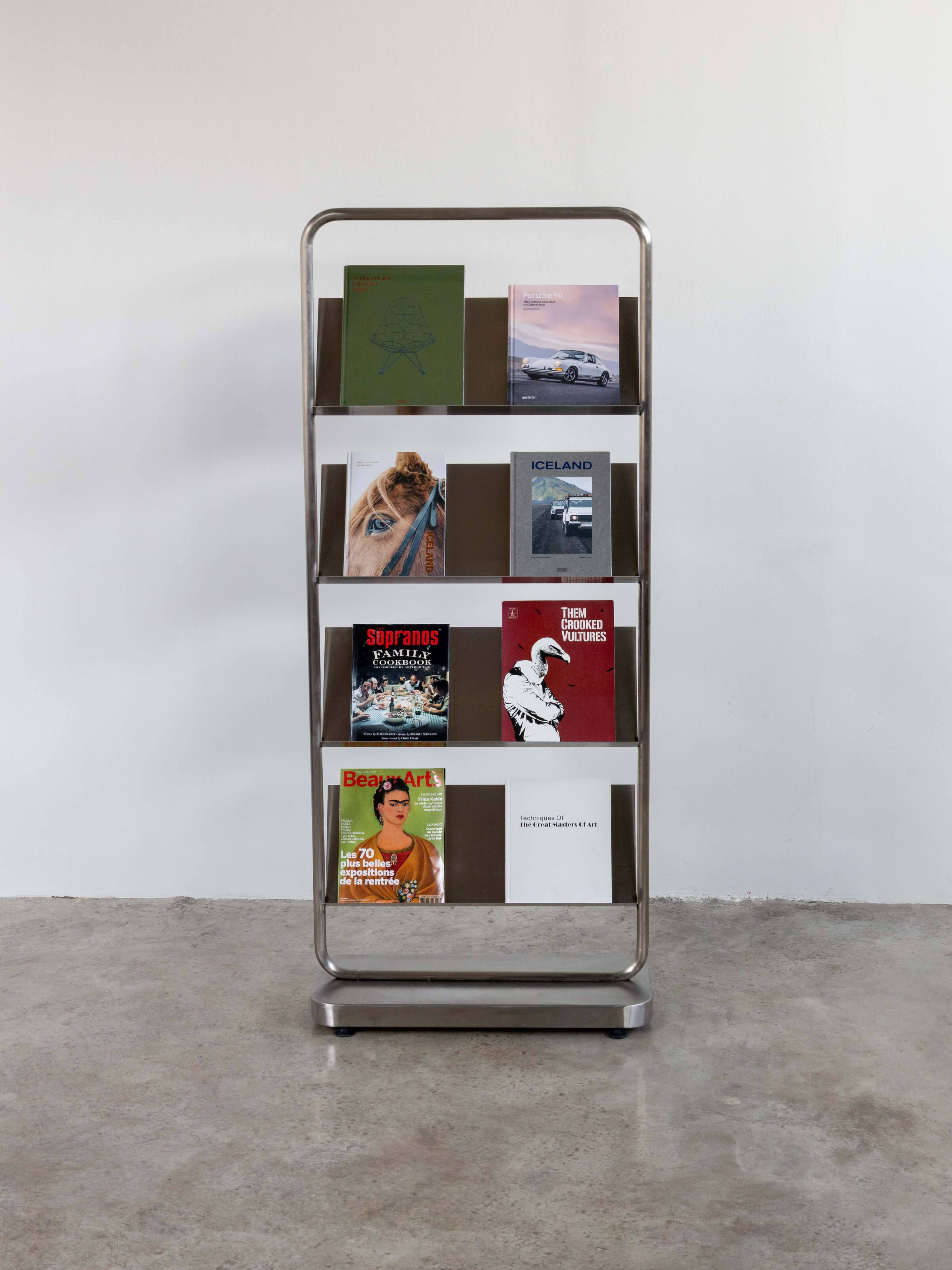 Stainless steel magazine rack with books on a concrete floor against a white wall