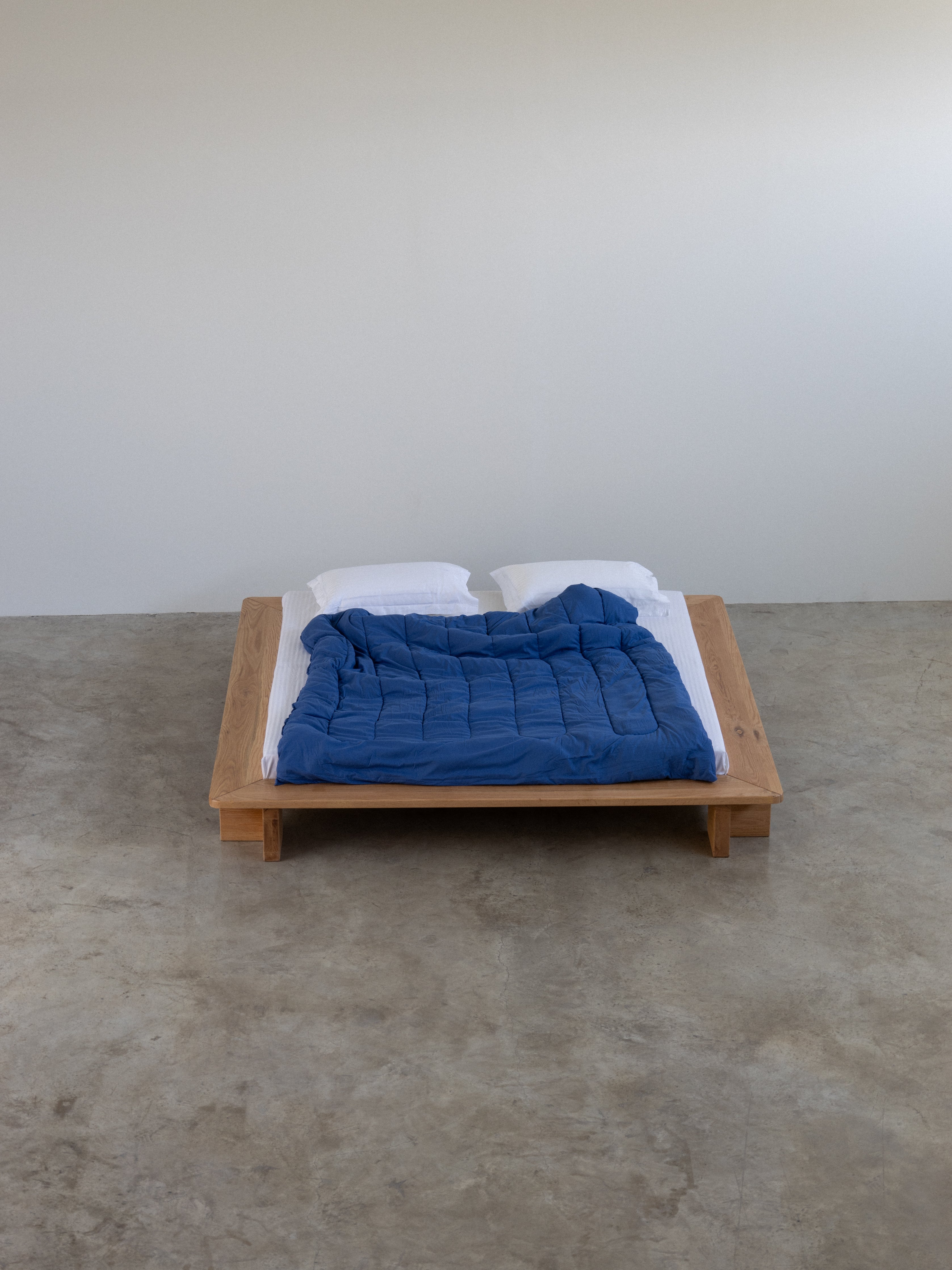 Wooden bed with blue blanket and white pillows on a concrete floor against a plain wall.