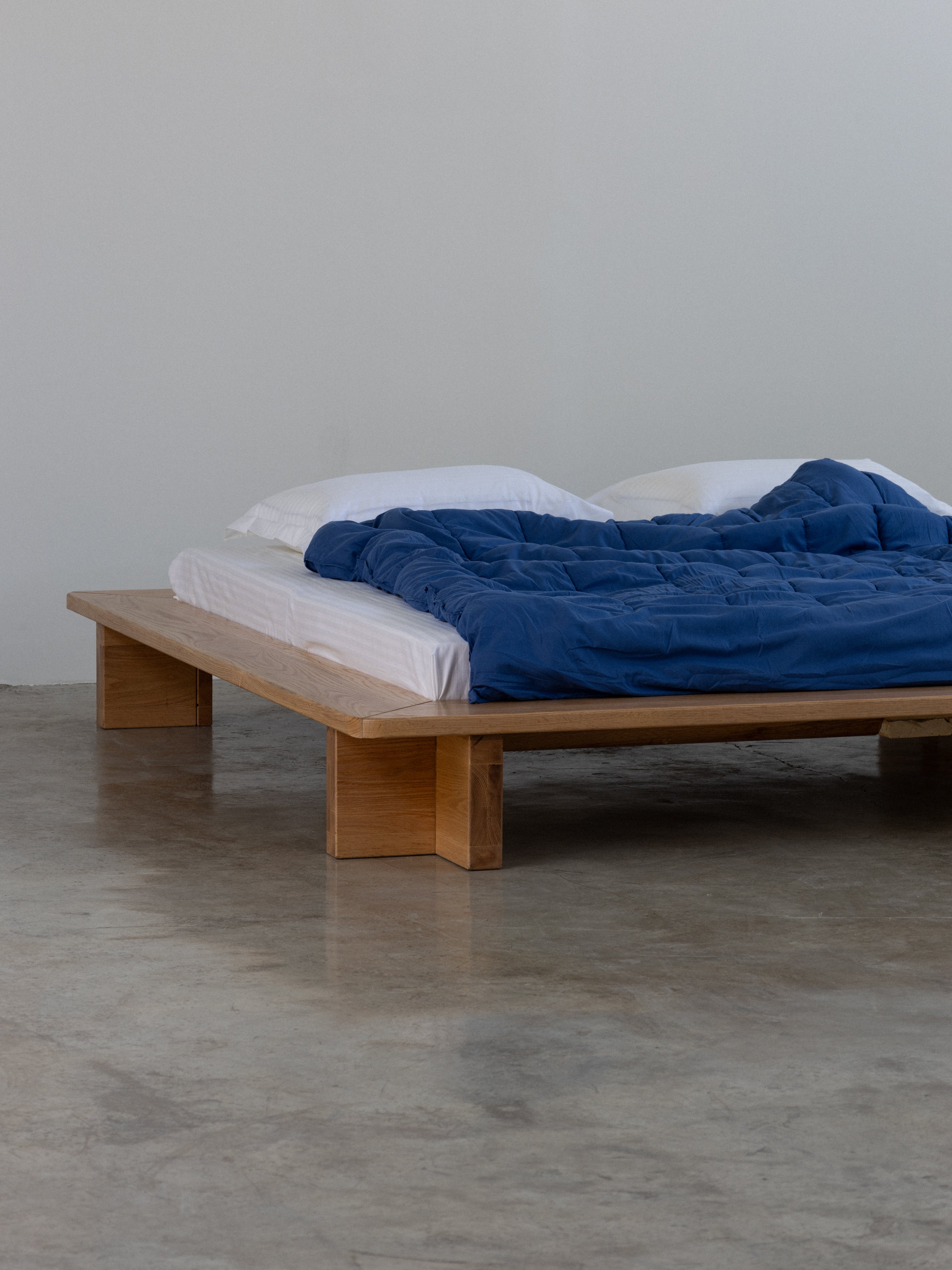A simple oak bed frame with a blue blanket and white pillows on it, placed on a concrete floor.