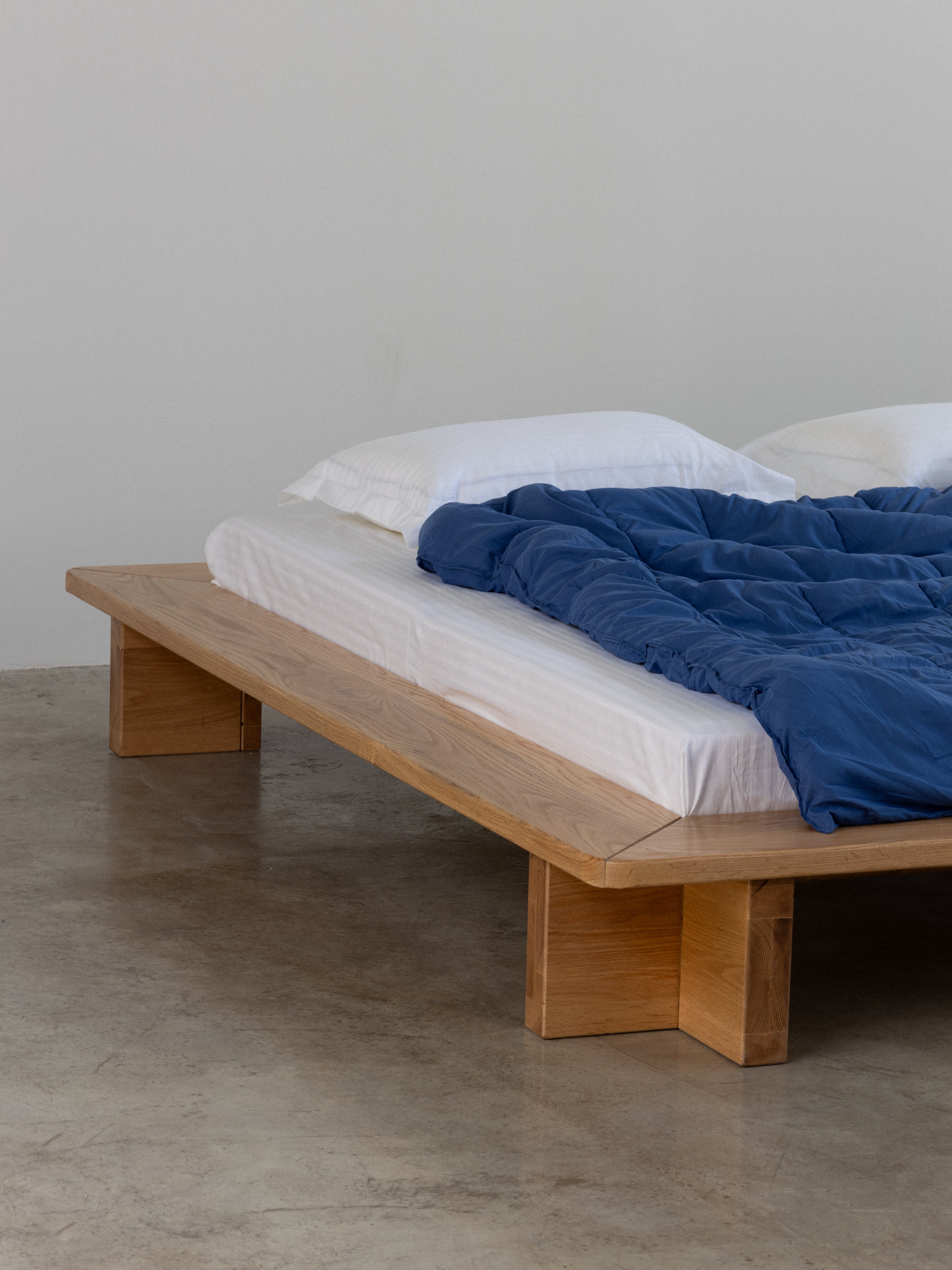 Wooden bed with white mattress and blue blanket against a plain wall.