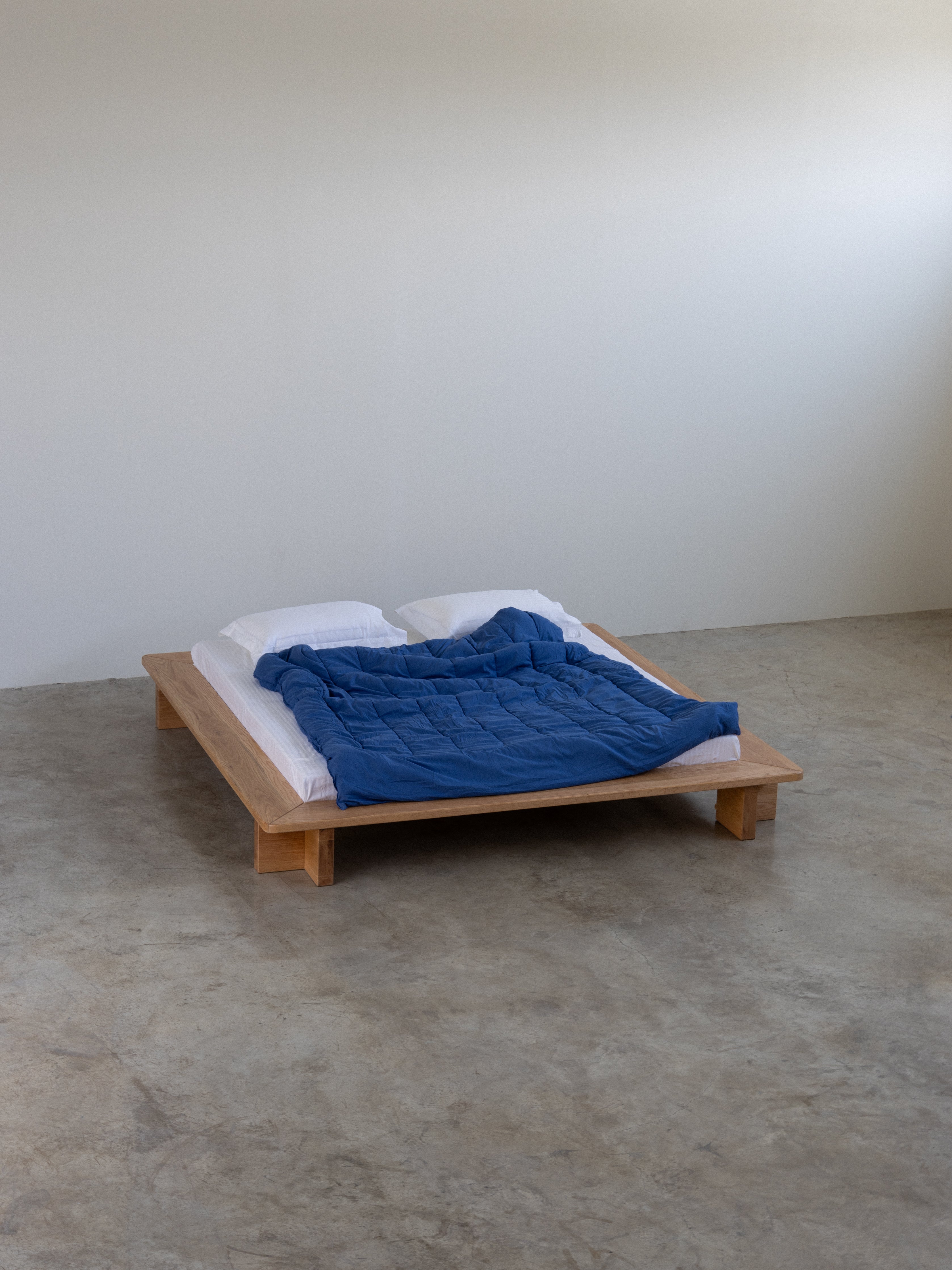 Wooden bed with blue blanket and white pillows on a concrete floor.