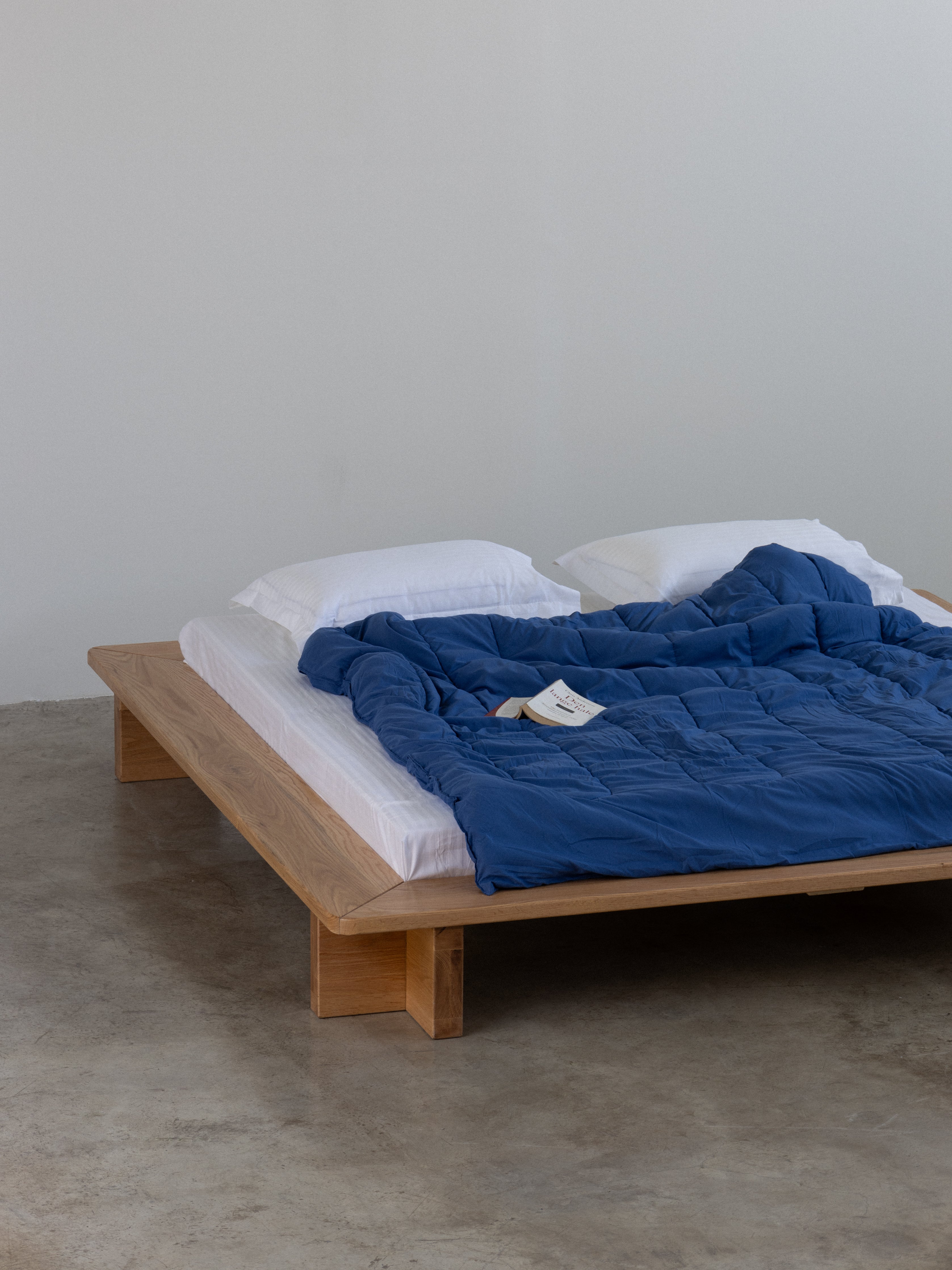 Wooden bed with white mattress and blue blanket against a plain wall.